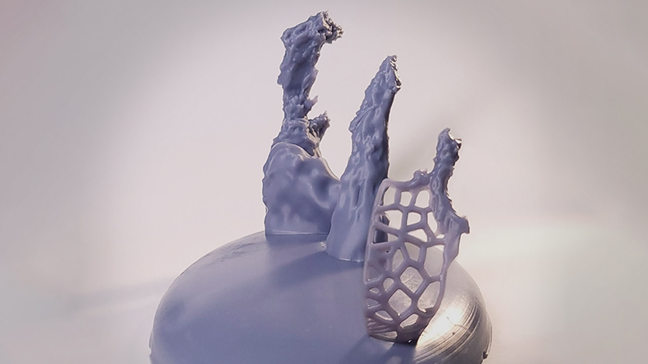 Photograph of a 3D printed model of the Pillars of Creation. The model is light gray with a silk-like texture, and the background is white. The three pillars rise up from a base that has a pancake-dome shape. The base is rotated about 60 degrees clockwise from the typical left-to-right orientation: The tallest pillar is at the back left at about 11 o’clock; the shortest is in the foreground right, at about 5 o’clock. The model is lit from several different angles to highlight the details. The pillars cast faint shadows toward the lower left. The pillars are craggy and stalagmite-like. The surfaces are smooth in some places, and pitted and grooved in others. Some edges are rounded and some appear sharp and jagged. The tall pillar at the back left and the middle pillar are solid all the way to the base. The small pillar at the front right is connected to the base by a web-like lattice made out of the same material as the rest of the model.