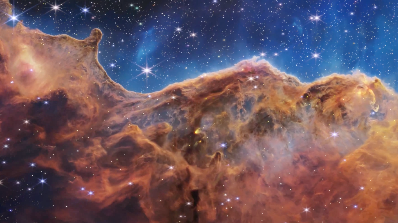 The image is divided horizontally by an undulating line between a brown cloudscape forming a nebula along the bottom and a comparatively clear upper portion in blue. Speckled across both portions is a starfield, showing innumerable stars of many sizes. The upper blue portion has wispy translucent cloud-like streaks rising from the nebula below. The orange and brown cloudy formation in the bottom half varies in density and ranges from translucent to opaque. The nebula contains ridges, peaks, and valleys—an appearance similar to a mountain range. In the bottom left corner, a clearer area free of gas and dust appears black with speckled stars.
